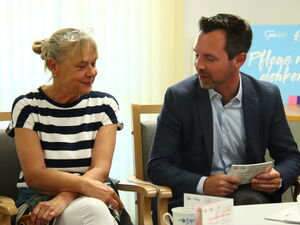 Frau mit blonden hochgesteckten Haaren und gestreiftem T-Shirt sitzt neben Mann in Anzug und Hemd, sie unterhalten sich.