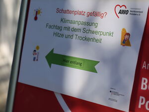Ein Schild mit einem grünen Pfeil nach links. Text: Schattenplatz gefällig? - Klimaanpassung - Fachtag mit Schwerpunkt: Trockenheit und Hitze