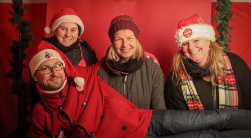 Drei Frauen lächeln, ein Mann liegt über deren queer auf ihren Beinen. Sie haben rote Weihnachmützen auf. Der Mann trägt eine Brille. Sie posieren vor einem roten Hintergrund.