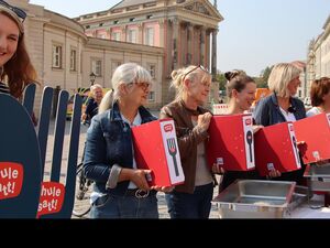 Auf dem Platz vor dem Landtag Brandenburg. 6 Frauen stehen nebeneinander. 5 von ihnen halten rote Aktenordner mit dem Schule Satt! Logo darauf.       Eine Frau hält große symbolisches Besteck Löffel und Gabel.