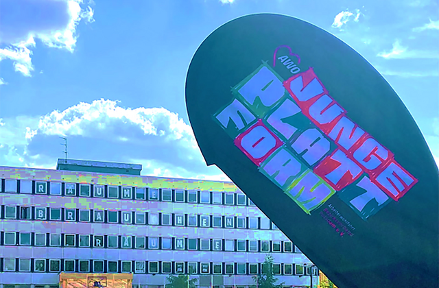 Gebäude, Rechenzentrum. Davor ein großer Sportplatz mit Basketballkorb und Toren. Die Sonne scheint. Wenige Personen sind auf dem Sportplatz. Werbefahne mit Aufdruck: AWO JUNGE PLATTFORM im Vordergrund.