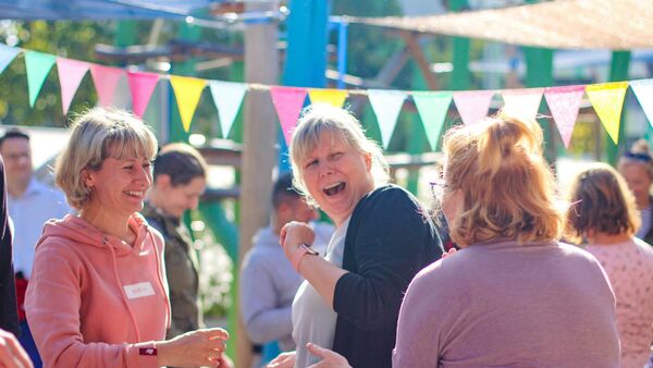 3 Frauen albern lachend herum. Draußen, Sonne scheint. Bunte Wimpel sind aufgehängt.