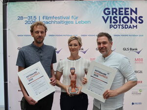 3 Personen vor Plakat von Green Visions Potsdam. Die Männer außen mit Urkunden in den Händen. Blonde Frau in der Mitte mit Skulptur.
