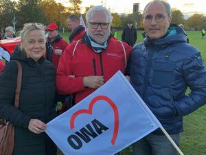 3 Personen, draußen auf einer Wiese. Blonde Frau trägt die Brille auf den Kopf geschoben mit schwarzer langer Jacke. Älterer Mann mit weißen Haaren, Brille und roter Jacke. Mann mit grauen Haaren, Brille, dunkelblauer Jacke. Eine weiße AWO Fahne.