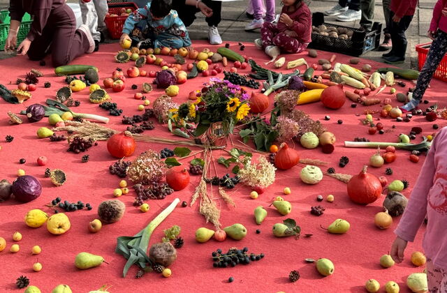 Großer runder roter Teppich darauf Blumen, Obst und Gemüse kreisförmig. Kinder und Erwachsene am Rand.