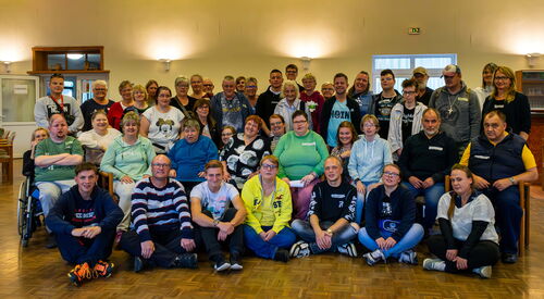 Großes Gruppenfoto in einer großen Halle. Vorne sitzen Menschen auf dem Fußboden, dahinter ist eine Reihe Stühle, ein Rollstuhlfahrer steht auch außen, dahinter stehen noch viele weitere Menschen.