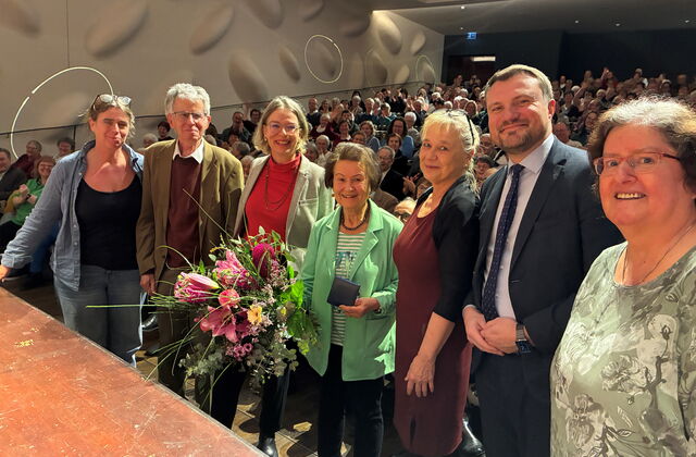 Preisverleihung: Gruppenbild vor der Bühne stehen 7 Menschen. Das Publikum hinter ihnen. Frau in der Mitte hält großen Blumenstrauß.