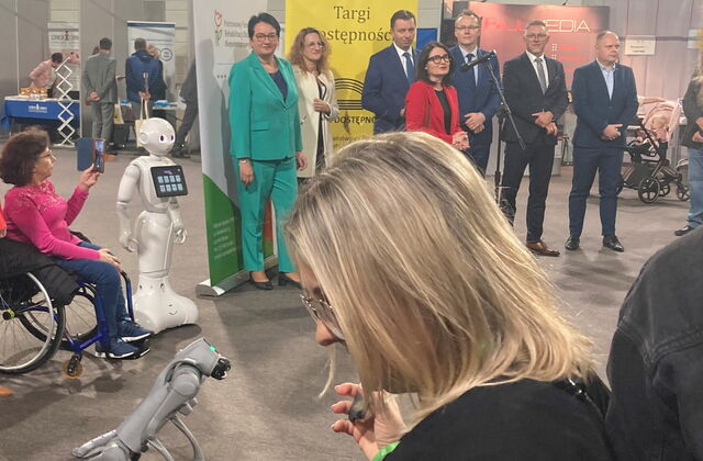 Ein weißer Roboter mit Armen, Gesicht und ein Touchpad steht neben einer Rollstuhlfahrerin im Aktivrollstuhl. Sie filmt mit ihren Smartphone die Rednerin. Hinter der Rednerin stehen noch zwei Frauen in Hosenanzügen und viel Männer in Anzügen.