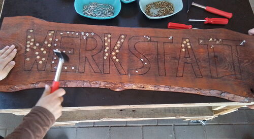 Großes Holzschild auf einer Werkbank. Auf dem Schild steht in Großbuchstaben das Wort Werkstatt.      Kinder füllen die Buchstaben mit Reiszwecken, Kind mit Hammer links im Bild.