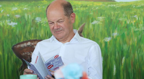 Mann sitzt vor einem Wandbild, dass eine weite Wiese mit Blumen zeigt. Mann guckt sich den Flyer von Kultur für Jeden an. Der Mann ist älter, hat eine Halbglatze, Haare an den Seiten sind kurz und grau. Er trägt ein weißes Hemd vor ihm eine Tasse.