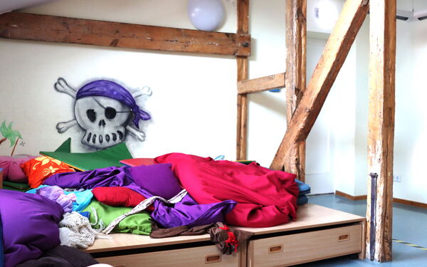 Holzbalken stützen den Raum. Eine Lümmelecke mit vielen farbigen Kissen und Decken, an der Wand dort ein Piraten-Totenkopf mit lila Kopftuch.