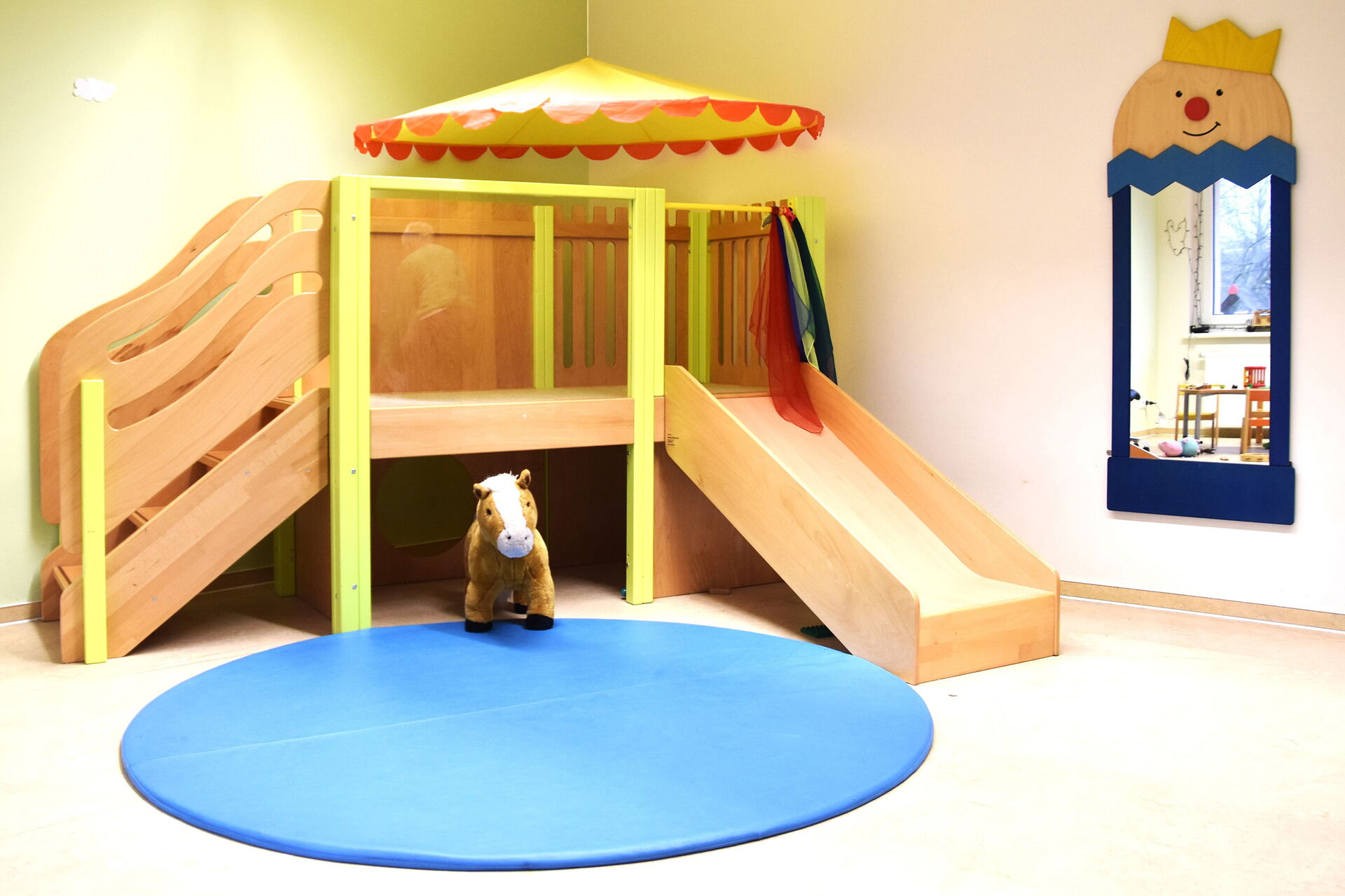 Helles hölzernes Klettergerüst mit einer Treppe und einer Rutsche, einem kleinen, auf dem Boden stehenden Pony-Kuscheltier und einem runden blauen Teppich.