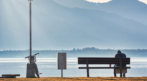 Eine Person sitzt einsam auf einer Bank am Wasser. Links neben der Bank ein Mülleimer noch weiter links lehnt ein Fahrrad an einer Laterne.