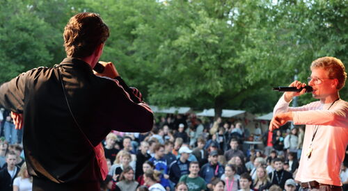 Blick von der Bühne ins große Publikum, zwei Männer mit Mikros auf der Bühne.