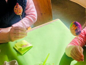 Beim Eier bemalen. Bunte Eier sind zum Trocknen auf Spieße gespießt. Eine Frau und ein Kind halten ihre Eier auf dem Spieß.