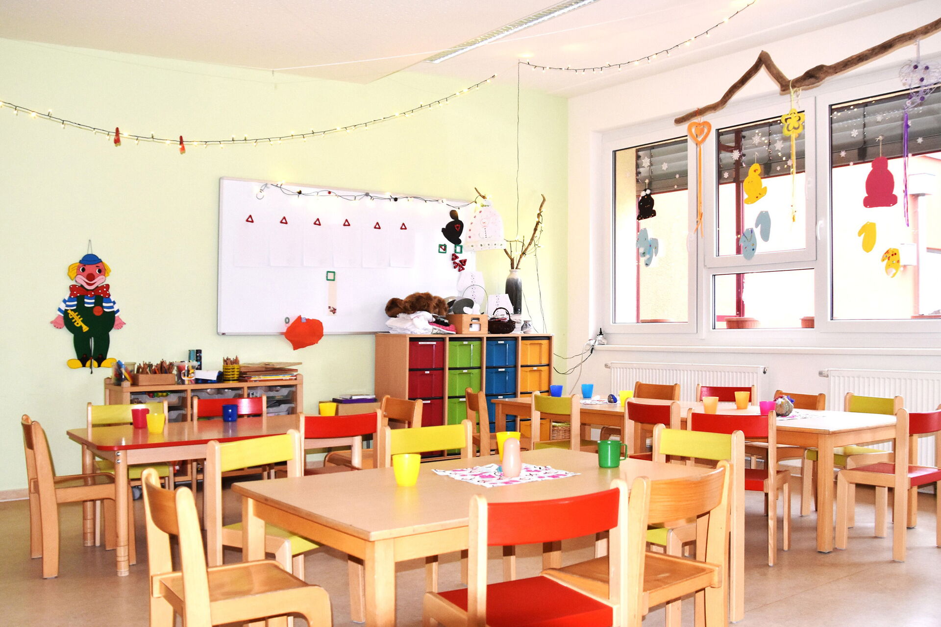 helleR, freundlicher Kindergartenraum. Im Vordergrund stehen mehrere kleine Tische mit bunten Kinderstühlen. An der Wand befindet sich eine große weiße Tafel, und ein farbenfroher Clown ist aufgehängt