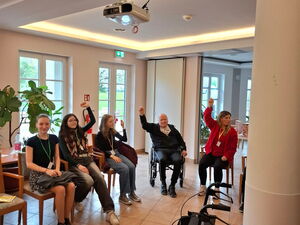 3 Schülerinnen und zwei Senioren sitzen auf Stühlen in einem Raum. Bewegungsübungen. Ein Arm nach oben.