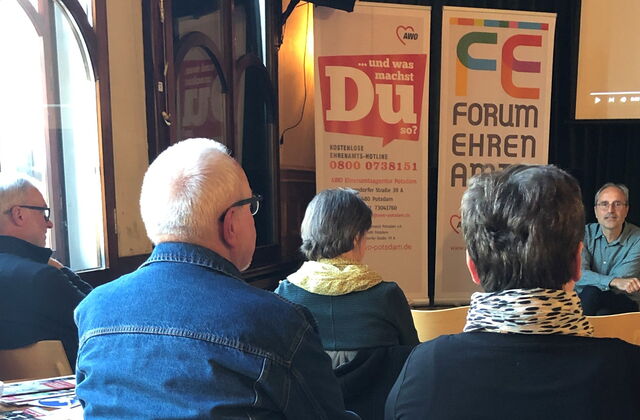 Das Bild zeigt mehrere Menschen im Zuschauerraum, die einem Mann zuhören, der auf der Bühne sitzt.