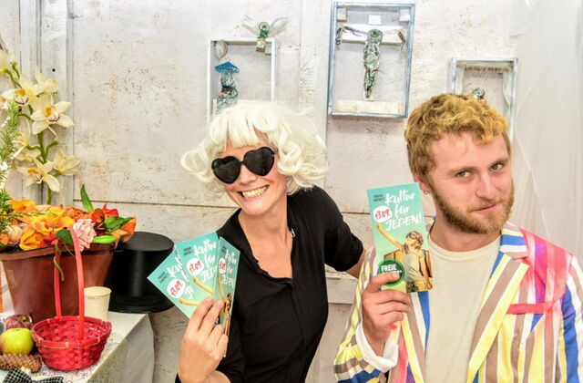 Zwei Menschen, Frau und Mann. Frau mit blonder wilder Frisur und Herz-Sonnenbrille hält drei Flyer von Kultur für Jeden in der Hand. Mann dunkelblond mit Bart einen Flyer von Kultur für Jeden in der Hand.