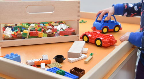 Spieletisch mit Lego und keinem Auto. Eine Kinderhand schiebt das kleine blaue Auto.