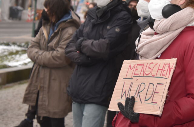 Draußen. Winter. Menschen mit FFP2 Masken in warmer Kleidung. Ein Mensch mit roter Jacke, schwarzen Handschuhen, und hellem Schal hält ein Pappschild vor die Brust. TextMENSCHENWÜRDE