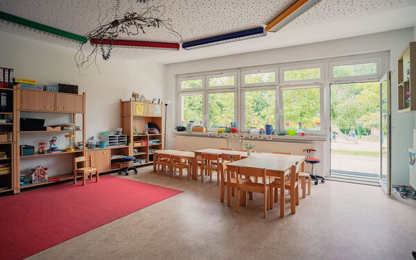 Großer heller Raum, die Terrassentür steht offen. Im Raum Tische mit Stühle, Regale mit Spielzeug an der Wand.