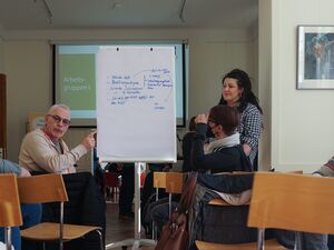 beschriebener Flipchart. Darum Sitzen Menschen aus der kleinen Gruppe und diskutieren ein Thema. Frau mit langen vollen schwarzen Haaren steht neben dem Flipchart.