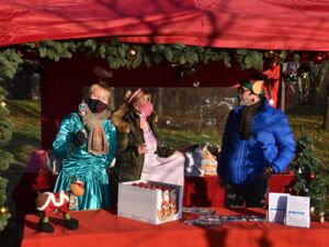 Ein weihnachtlich geschmückter Stand, Eine Kiste Schoko-Weihnachtsmänner und ein Rudolf-Rentier stehen auf dem Tisch. Im Stand zwei Frauen als Engel und Fee verkleidet und ein Mann in blauer Winterjacke.