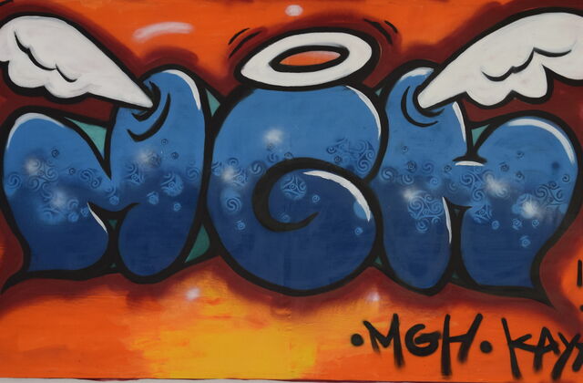 Graffiti: Blaue Buchstaben auf oragenen Hintergrund. MGH       über dem G ist ein Heiligenschein, und die anderen Buchstaben haben Engelsflügel.