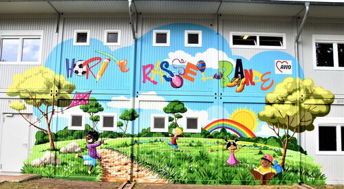 Wandbild an Fassade eines Gebäudes. Bunte Schrift: Hort Rasselbande. Spielende Kinder, Regenbogen, Sonne, Wiese Bäume