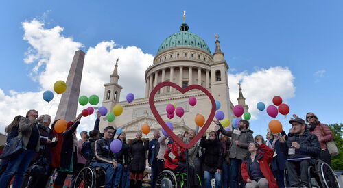 Vor der Nikolaikirche Potsdam, VViele Menschen mit und ohne Behinderung, u.a. drei Rollstuhlfahrer. In der Mitte ist ein großes Herz aufgestellt, und die Menschen lassen bunte Luftballons fliegen.