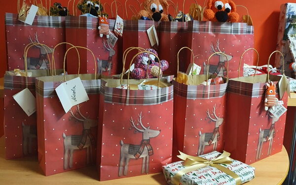 Rote Geschenktüten mit Rentier drauf, gut gefüllt stehen auf einem Tisch. Davor auf Tisch noch ein kleines Päckchen mit weißem Papier und goldenem Schleifenband.