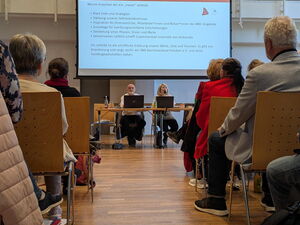 Gang zwischen Stuhlreihen, Vorne sitzen zwei Menschen an Laptops. An einer Leinwand über ihnen ist Text projiziert.
