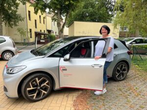 Ein silbernes VW Auto mit dem Logo der AWO Sozialstation auf der Fahrertür. Die Tür ist geöffnet und eine Frau mit schwarzen vollen Haaren, einem hellen T-Shirt und blauer dreiviertel-Jeans steht an der Fahrertür und und lächelt in die Kamera.