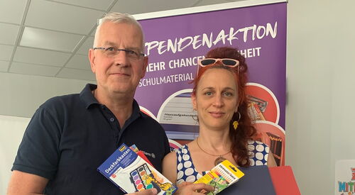 Mann und Frau stehen nebeneinander im Büro. Beide halten Schulmaterial in den Händen, Hefter, Stifte, Tuschkasten. Mann hat graue kurze Haare, Brille und ein dunkelblaues Poloshirt. Frau hat lange rote Haare, Brille auf den Kopf geschoben in trägt Kleid.