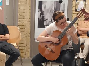 Frau mit Gitarre sitz auf Stuhl in einem Raum. An den Wänden des aus sind Bilder, Gemälde. Frau trägt, helles T-Shirt, Jeans, Turnschuhe und ein braunes Haarband sowie Brille, Sie spielt auf der Gitarre.