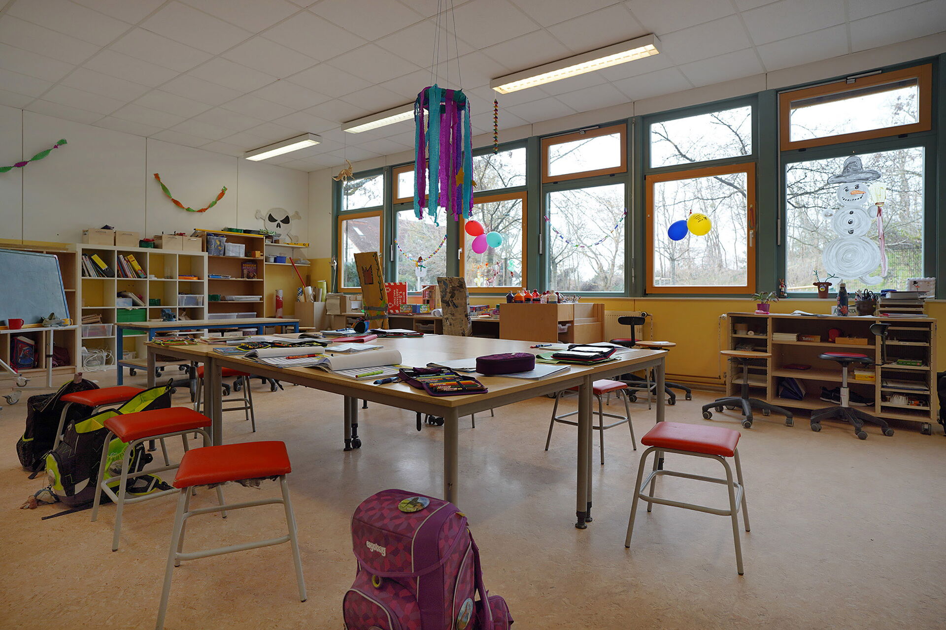 Lernzimmers. großer Tisch in der Mitte des Raumes, darauf Federtaschen, Blöcke und Hefter. Um den Tisch stehen Hocker und Schulranzen. An den Wänden stehen Regale.