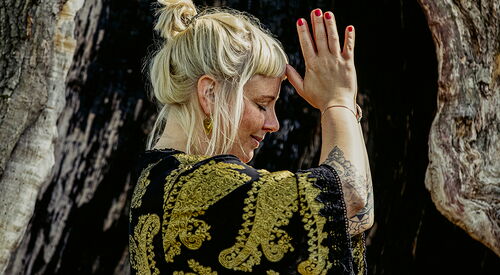Blonde Frau in lockerer Kleidung. Sie steh seitlich und hat die Hände an einander auf Kopfhöhe - ihr Gesicht wirkt entspannt.