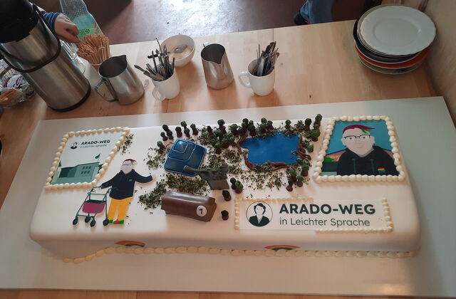 Eine rechteckige Torte mit weißem Zuckerguss ist verziert mit Figuren, Pyramide, Schokoriegel und Flächen mit schwarzer Schrift: Arado-Weg in Leichter Sprache