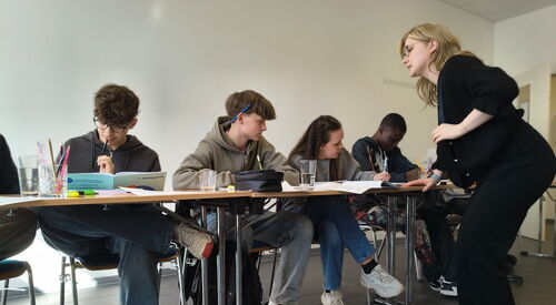 4 Jugendliche sitzen an aneinandergereihten Tischen nebeneinander. Auf den Tischen Schulmaterial. Ihnen gegenüber auf der innenseite des Tisches eine blonde Jugendliche stehend.
