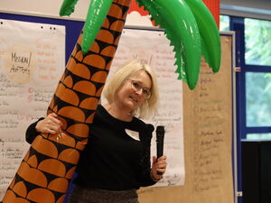 Innenraum. An der Wand stehen beschriebene Flipchart-Wände. Im Vordergrund umarmt eine blonde Frau mit Mikro in der Hand eine Luft-gefüllte Gummi Palme.