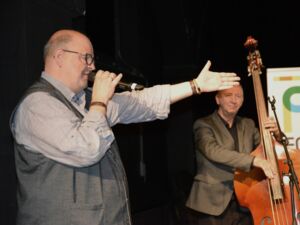 Älterer Mann mit Mikrofon in der Hand weißt mit dem anderem Arm auf die Band Mitglieder. Mann trägt Brille, hellgraues Hemd und eine dunkelgraue Weste.