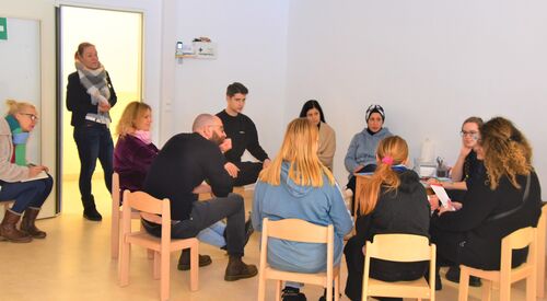 Menschen unterschiedlicher Nationalitäten sitzen in einem Raum im Kreis. Eine Frau steht in der offenen Tür.