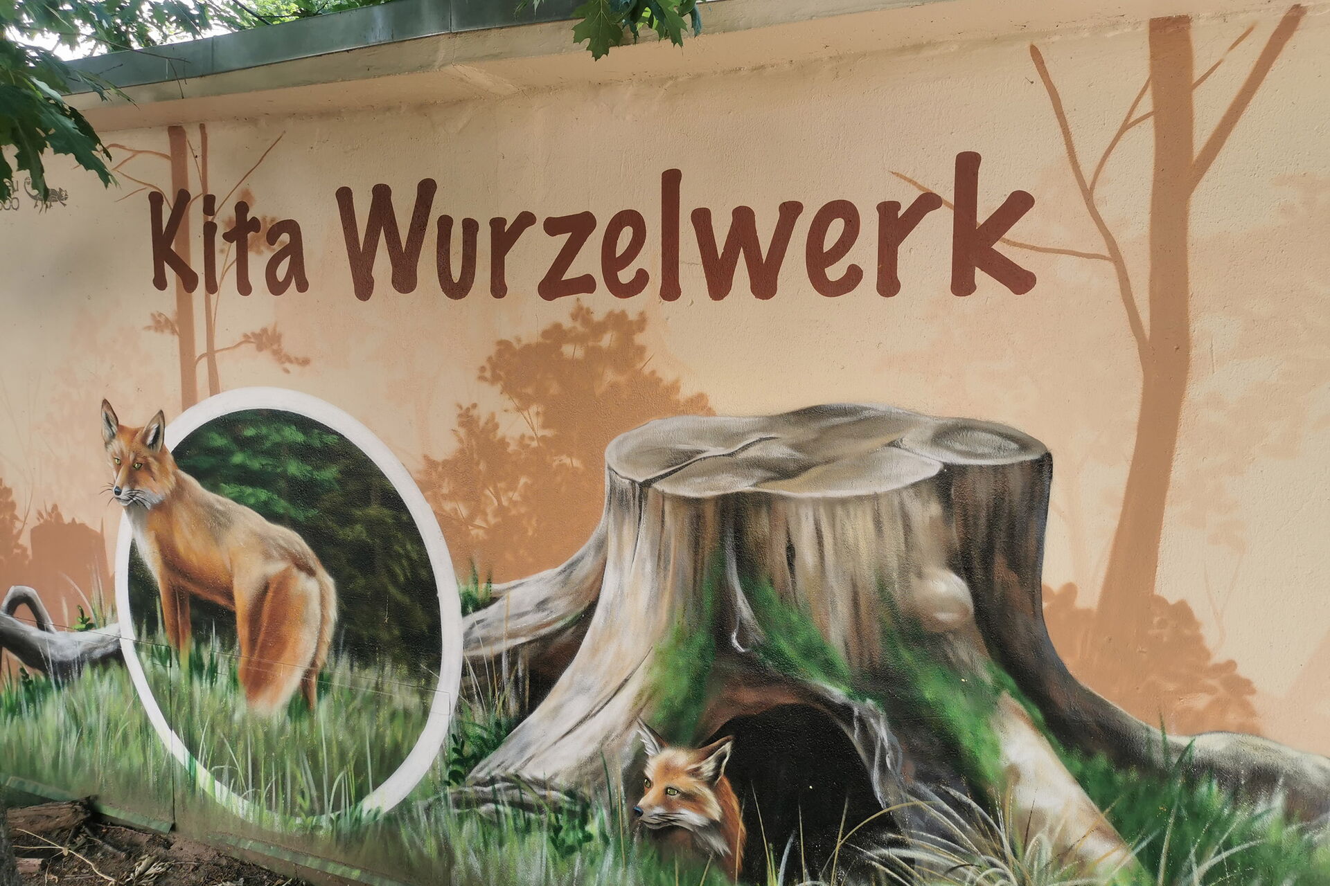 Eine Häuserwand mit einem Wandbild von eine Fuchsbau unter einem großen Baumstumpf. Ein Fuchs schaut aus dem Bau. Drüber steht groß: Kita Wurzelwerk