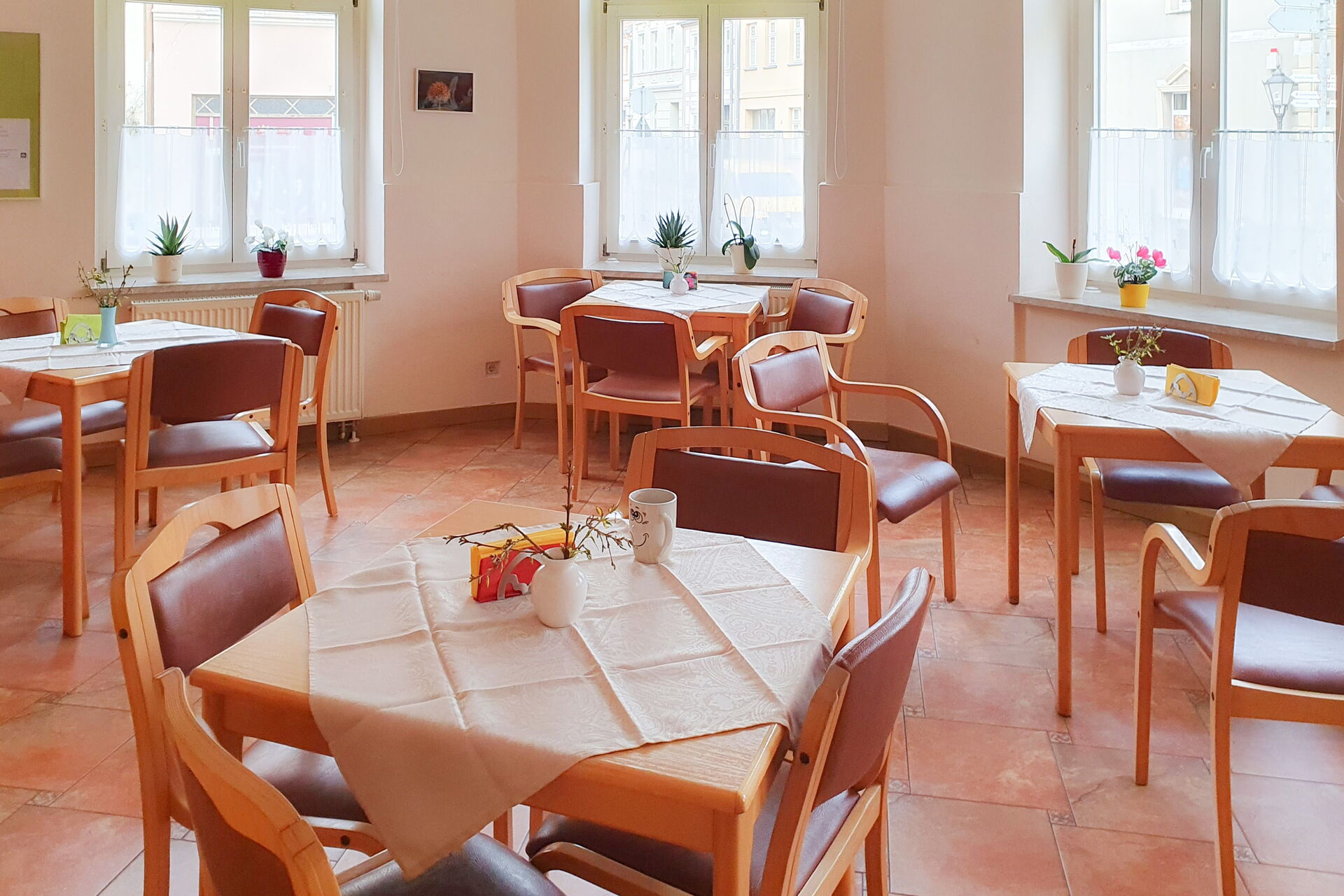 Ein Speisesaal. 4 kleine vierer Tische im Raum verteilt. Helle Raum, Fenster lassen viel Licht herein.