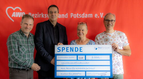 Vier Personen vor einer roten Wand mit AWO Logo und Schriftzug. Eine Frau und ein Mann halten einen großen Scheck in die Kamera. Zwei weitere Männer stehen links daneben. Alle vier lächeln in die Kamera.