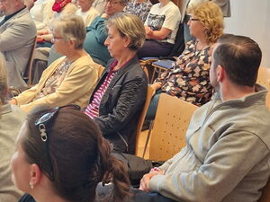 Menschen sitzen auf Stühlen in einem Raum. Hinten steht eine Frau mit Mikrofon. Einige drehen sich zu der Frau mit Mikro um.