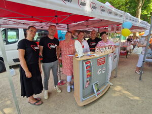6 Personen, darunter ein Mann, stehen an einem Stand mit einem klein Glücksrad und lächeln in die Kamera.