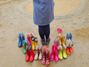 Bei einem Spielplatz. Eine Person mit Rücken zur Kamera hält einen bunten Kinderregenschirm. Um die Person stehen mehrere Paare bunter Kinderstiefel.