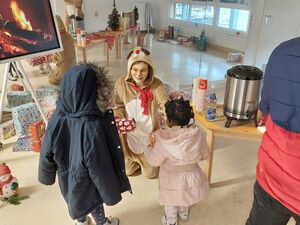 Großer heller Raum. Großer Fernseher  mit Lagerfeuer-Bild. Davor kniet eine Person im Rentierkostüm und übergibt Geschenke an zwei kleine Kinder.